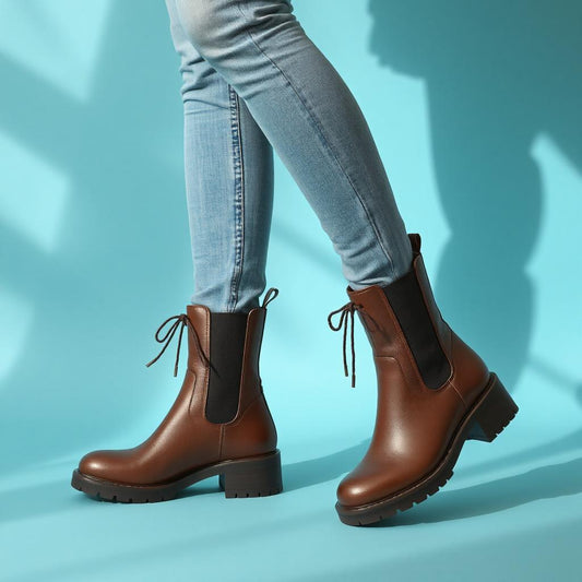 Woman wearing brown Chelsea boots with mid block heel and short front laces, paired with blue jeans, on a blue background.