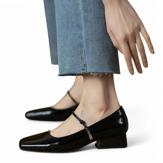 Black shoe beside model’s hand, showing flared block heel and tiny gold-tipped strap end.