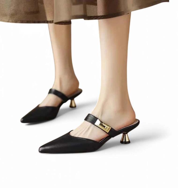 A pair of black pointed-toe mules with gold trumpet heels worn beneath a sheer chiffon skirt.