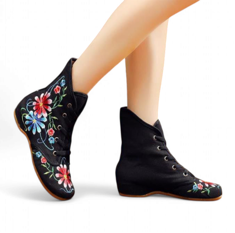 Black embroidered floral boot on a model’s foot, side angle showing low hidden wedge and lace-up front.

