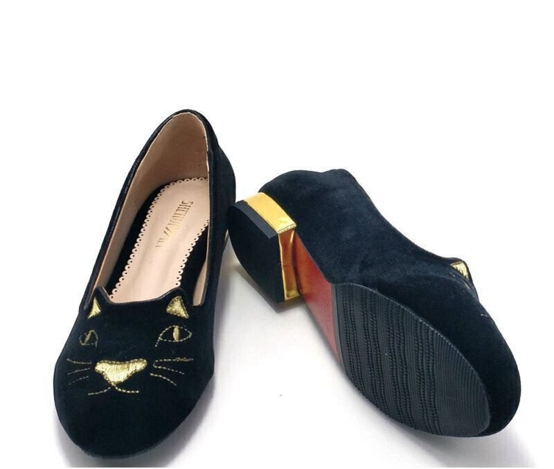 Three-quarter pair view showing black suede flats, cat face embroidery, rubber outsole and gold heel accent.