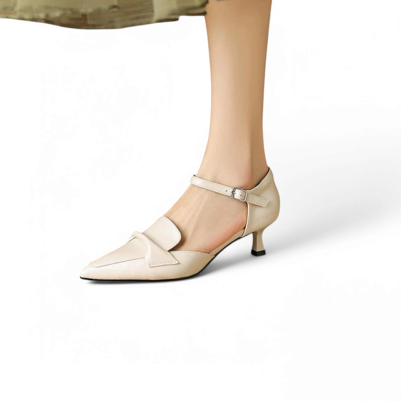 Cream cow-leather pump with pointed toe, metal ankle buckle and low kitten heel, shown on foot with a skirt.