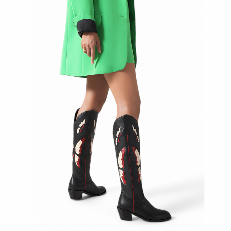 Side view of black boot with red butterflies and red piping as model walks.

