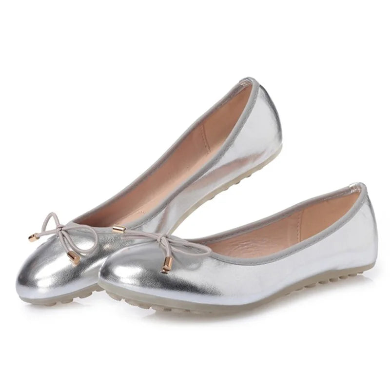 Pair of silver metallic round-toe ballet flats with small bow and dot-grip rubber outsole on a white background; front-angled view
