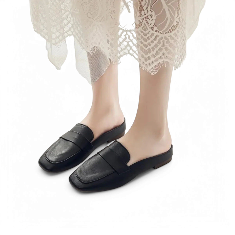 Angled front/side view of the black Merendy Snow square-toe flat mule worn with a lace hem dress; open back.
