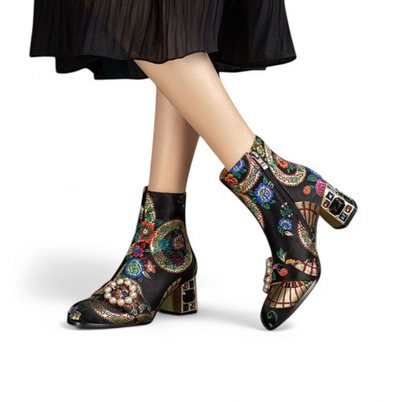Black embroidered ankle-boot pair with pearl buckle and jeweled block heels, worn with a pleated skirt.